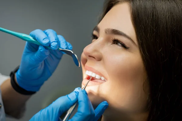 Leende glad brunett kvinna patient granskas av tandläkare i blå handskar med hjälp av tandspegel och scaler sitter en halv sväng i tandstol, sjukvård koncept — Stockfoto