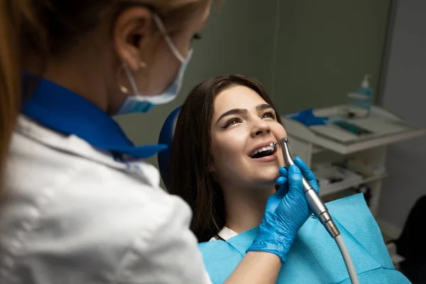 Brunetka žena pacient vyšetřen zubař v modrých rukavicích a masce pomocí zubního zrcadla a škálovač sedí v zubním křesle během vytvrzování zubů na klinice, helthcare koncept — Stock fotografie