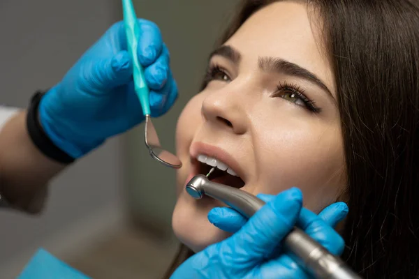 Dentist wearing blue gloves examines smiling woman patient's oral cavity using dental scaler and mirror while she sits in dental chair in clinic , healthcare concept — Stock Photo, Image