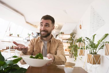 Genç gülümseyen adam salatayla tabağı tutuyor ve öğle yemeğini kafede yiyor. .