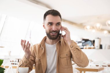 Genç yakışıklı adam, kafede kahve içiyor ve akıllı telefonuyla konuşuyor, birden fazla işi var..
