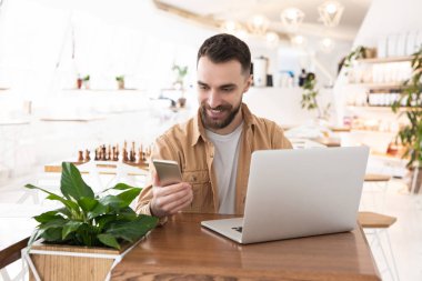 Laptopunda çalışan yakışıklı bir genç kafeterya molasında akıllı telefon kullanıyor, öğle yemeği zamanı, birden fazla görev konsepti..