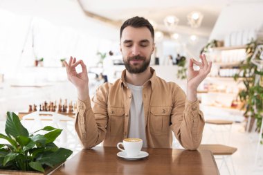 Genç sakallı adam yemek molasında kahve içmeden önce yoga pozisyonunda oturur, sakin bir konsept..