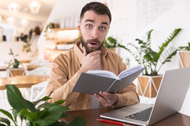 Genç ve yakışıklı sakallı bir adam öğle yemeği arasında kafede çalışırken dizüstü bilgisayarını kontrol ederken şaşkınlıkla bakıyor..