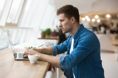 Genç yakışıklı adam öğle yemeğinde salata ve sıcak kahve içen dizüstü bilgisayarında ciddi görünüyor ve birden fazla işi olan bir kafeye odaklanmış..
