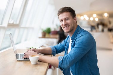 Genç, yakışıklı, gülümseyen adam laptopunda salata yiyip öğle yemeğinde sıcak kahve içiyor. Kafede otururken mutlu görünüyor..