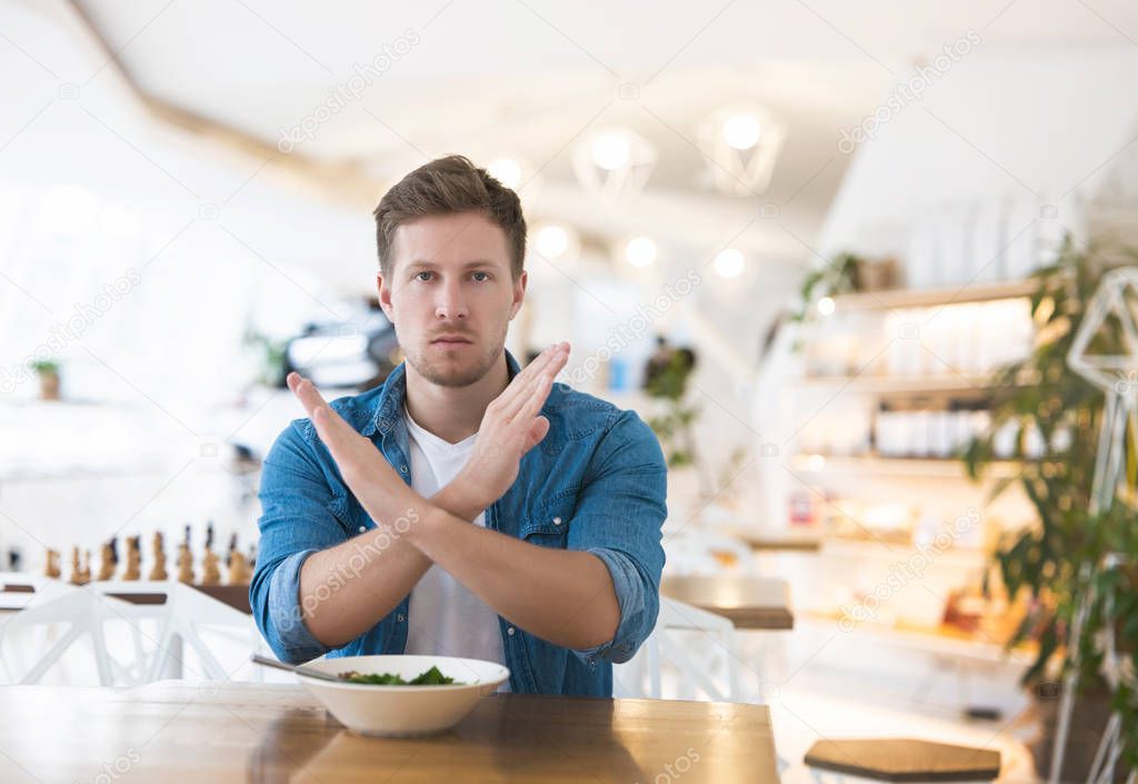 handsome man showing no sign crossing his arms while having lunch at cafe during break, refuses to meat, prefers eat salad, vegan and dietology concept.