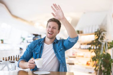 Genç yakışıklı, mutlu bir adam kafede otururken arkadaşına el sallıyor. Vücut dili kavramı..