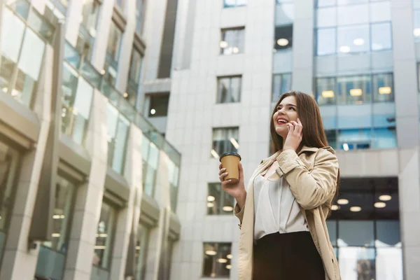 businesswoman drinking hot coffee on the go in the middle of the street standing on background of office center talking on her phone, multitasking concept.