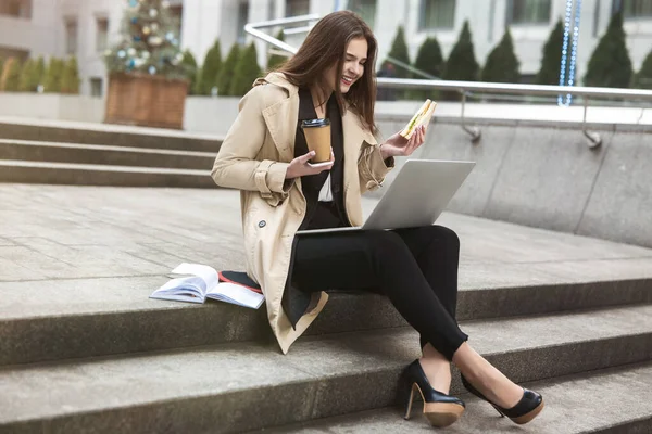 young beautiful businesswoman with sandwich and hot coffee talks online with her friend using laptop sitting on stairs outside office during lunch break, multitasking concept.
