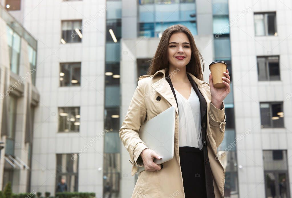 happy business lady holding laptop under arm walking down the street near office center drinks coffee in paper cup feeling positive, big city life concept.