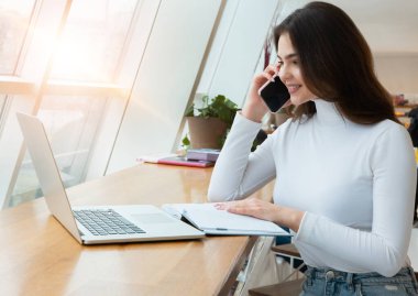 Genç ve güzel iş kadını laptopunda çalışıyor ve yemek molasında kafede otururken telefonla konuşuyor..