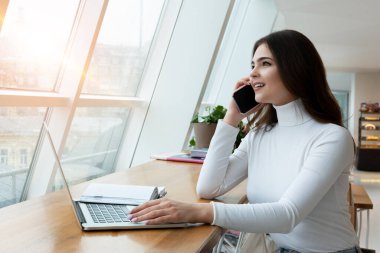 Genç ve güzel iş kadını, öğle yemeği molasında laptopunda çalışırken telefon görüşmesi yapıyor..
