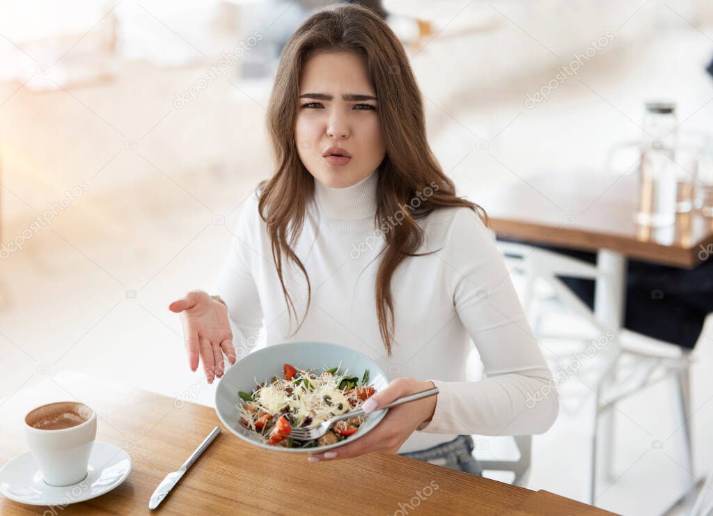 young beautiful woman looks unsatisfied with quality of salad holding plate looking unhappy during lunch time at cafe, , complaining guest concept.