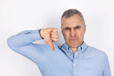 Gri saçlı, mavi tişörtlü yetişkin ve yakışıklı bir adamın portresi. İzole edilmiş beyaz arka planda duran işaretten hoşlanmadığını gösteriyor..