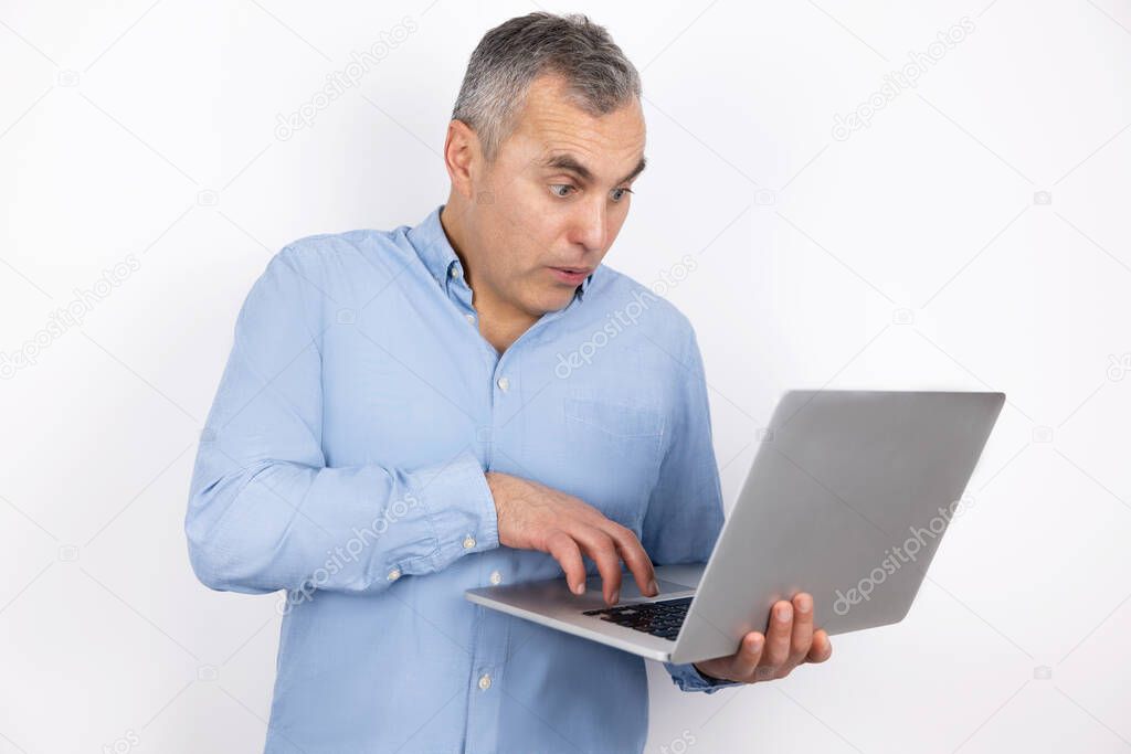adult handsome man with grey hair wearing blue shirt types on his laptop standing on isolated white background, modern technology concept.