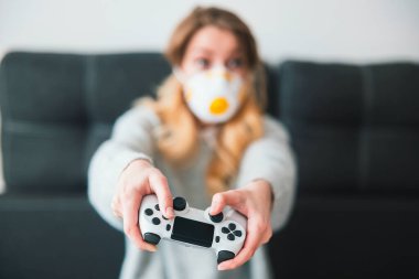 Tıbbi koruma maskesi takan genç sarışın kadın ellerinde joystick ile oyun oynuyor. Karantina sırasında evde oturuyor. Virüs salgını konsepti..