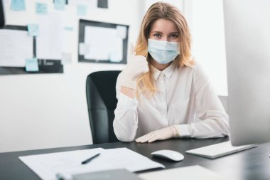 Tıbbi koruma maskesi ve eldiven giyen genç sarışın kadın yönetici Covid-19 epidemisi sırasında ofiste çalışıyor..