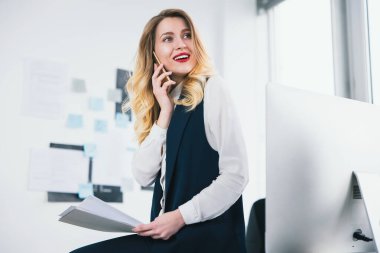 Genç bir kadın yönetici elinde belgelerle iş ortağıyla telefonda konuşurken meşgul görünüyor. Çoklu görev, iş kavramı..