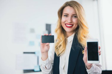 Genç, gülümseyen bir kadın yönetici, bir elinde banka kartıyla parlak ofisinde duruyor, diğer elinde akıllı telefonu, birden fazla işi olan, modern teknoloji kavramını gösteriyor..
