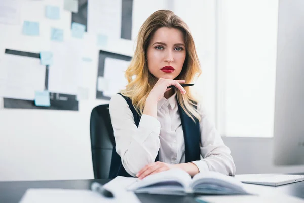 young blond beautiful woman manager with red lipstick works in her modern office, holding her hand with pen near face, looks sexy, multitasking, work concept.