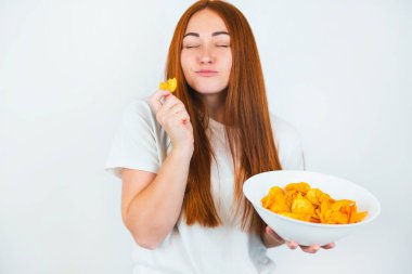 Kızıl saçlı, patates cipsi yiyen genç bir kadının portresi izole edilmiş beyaz arka planda, abur cubur konseptinde mutlu görünüyor..