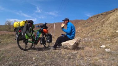 Kojomkul, Chuy Region, Kyrgyzstan - October 06, 2019: Bicycle traveler sits on a stone and eats a flatbread. A bicycle is standing nearby.