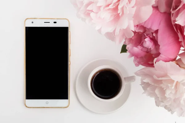 Coffee cup with phone and peonies on white background. Flat lay. Top view. — Stock Photo, Image