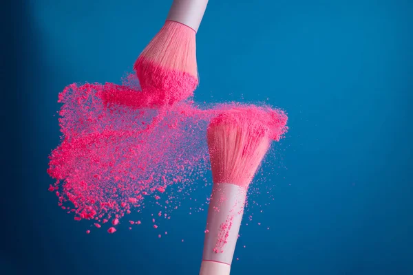 Foto de close-up de dois pincéis de maquiagem profissional com pó rosa em movimento no fundo azul — Fotografia de Stock