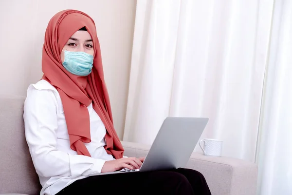 Korunmak için cerrahi maske takan Müslüman bir kız. Başörtülü kadın Coronavirus ya da COVID-19 için maske takıyor ve evinde oturuyor.
