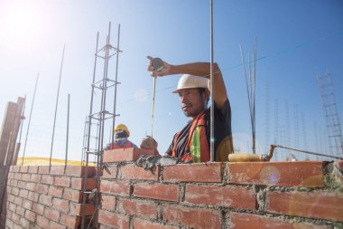 Meksikalı adam tuğla ve çimentoyla bir ev duvarı inşa ediyor. Kask ve yelek ile donatılmış.