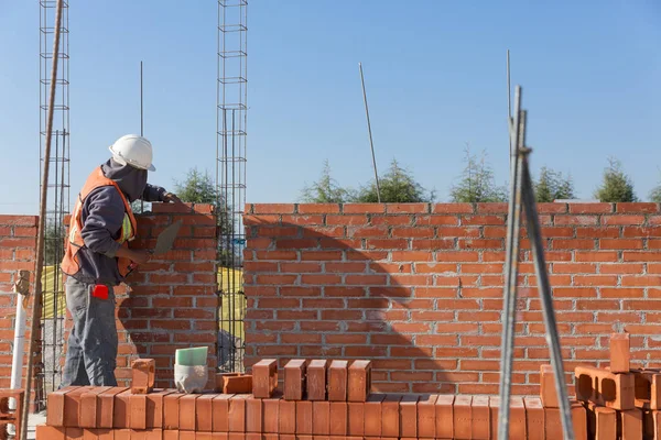 Meksikalı adam tuğla ve çimentoyla bir ev duvarı inşa ediyor. Kask ve yelek ile donatılmış.