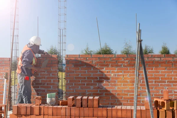 Meksikalı adam tuğla ve çimentoyla bir ev duvarı inşa ediyor. Kask ve yelek ile donatılmış.