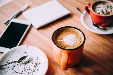 Bir fincan cappucino, akıllı telefon ve kafedeki ahşap bir masanın üzerinde defter.
