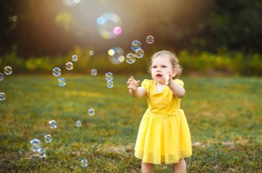 Yeşil glade içinde sarı elbiseli küçük bir kız sabun köpüğü yakalıyor.