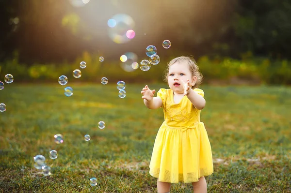 Yeşil glade içinde sarı elbiseli küçük bir kız sabun köpüğü yakalıyor.