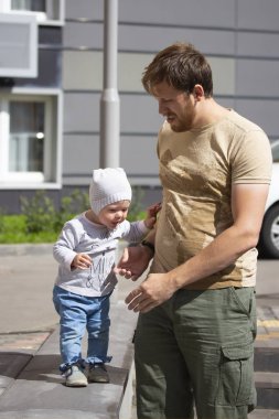 Genç bir baba çocuğuna yürümeyi öğretir. Küçük kız, kot pantolonlu ve şapkalı bir çocuk babasıyla yürüyor. Aile yürüyüş yapıyor, sıcak güneşli bir gün.