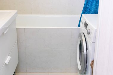 Beyaz mermer kiremitlerle dekore edilmiş küçük bir dairede dar bir banyo. Bir kaideyle kaplı, dökme demir bir küvet, gümüş varilli bir çamaşır makinesi.