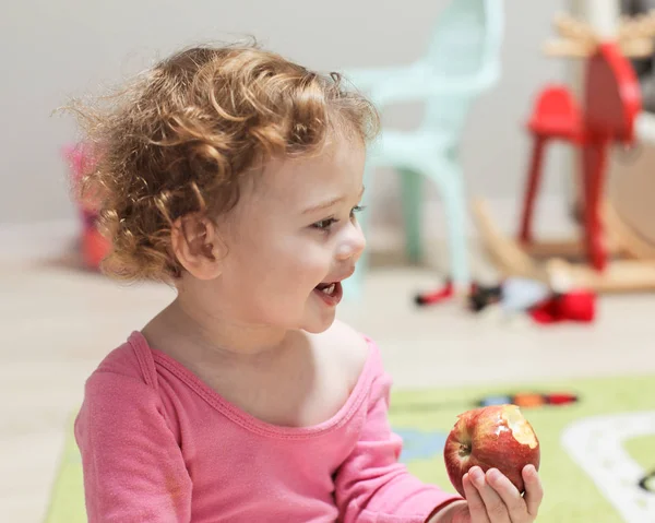 Kis fehér lány rózsaszín eszik egy almát, és mosolyog egy almát. Vidám boldog mosolygós baba göndör hajjal az otthoni belső térben, portré egy nevető kislányról — Stock Fotó