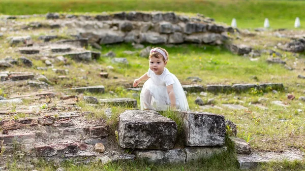 Little Caucasian girl in a white dress climbs on the old stones of the ruins. Beautiful elegant child walking in the summer on the meadow, baby girl outdoor — стокове фото