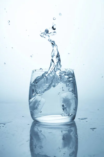 Mooie plons, ijsblokje dat in het water valt, een glas met schoon drinkwater en een hoge plons met waterdruppels — Stockfoto