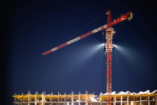 Construction crane in night illumination, construction of a multi-storey modern residential building. Night construction site, twilight, spotlight. Construction crane lighting