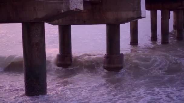 Les vagues de la mer se brisent sur le rivage rocheux et sur les supports du pont — Video