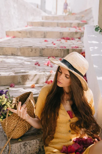 Fille dans une robe jaune et chapeau avec de longs cheveux bruns, est assis sur les marches, sur le fond de cactus à fleurs vertes et fleurs de bougainvilliers roses, en été en Méditerranée, en vacances — Photo