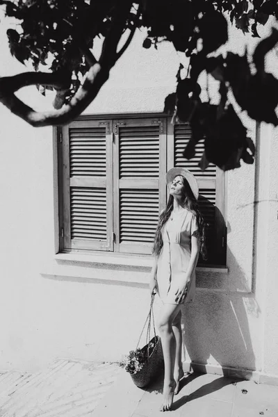 Chica en un vestido y sombrero con el pelo largo de pie junto a una casa bajo el árbol en verano en el Mediterráneo, de vacaciones —  Fotos de Stock