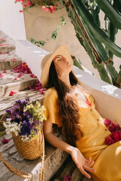 Fille dans une robe jaune et chapeau avec de longs cheveux bruns, est assis sur les marches, sur le fond de cactus à fleurs vertes et fleurs de bougainvilliers roses, en été en Méditerranée, en vacances — Photo