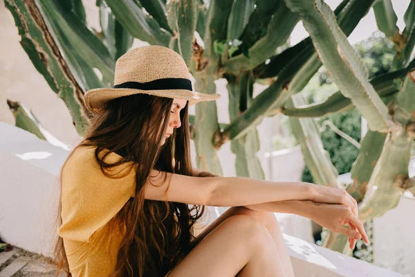 Chica en un vestido amarillo y sombrero con el pelo castaño largo, está sentado en las escaleras, en el fondo de cactus de floración verde y flores de buganvillas rosadas, en verano en el Mediterráneo, de vacaciones —  Fotos de Stock