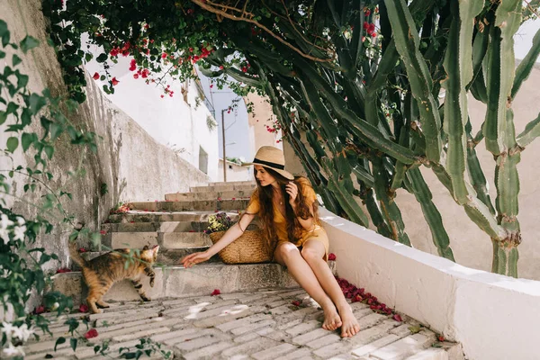 Sarı elbiseli ve şapkalı kız, merdivenlerde kedisiyle oturuyor, yeşil çiçekli kaktüs ve pembe bougainvillea çiçeklerinin yanında, Akdeniz 'de, tatilde.