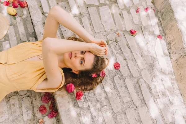 Chica en un vestido amarillo con el pelo castaño largo, se encuentra en las escaleras, vista superior en el fondo de flores de buganvillas rosadas, en verano en el Mediterráneo, de vacaciones —  Fotos de Stock