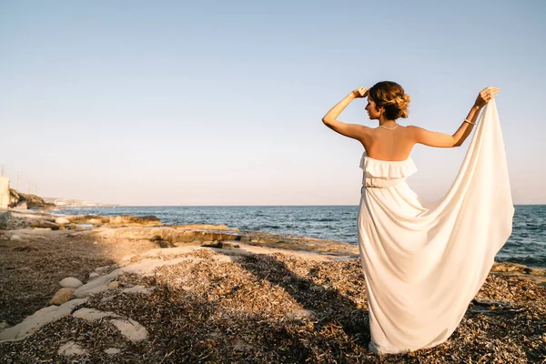 ギリシャ風の白いドレスを着た美しい若い笑顔の女の子が海の遊歩道を歩いています。夏時間日没時間。海での花嫁結婚式 — ストック写真